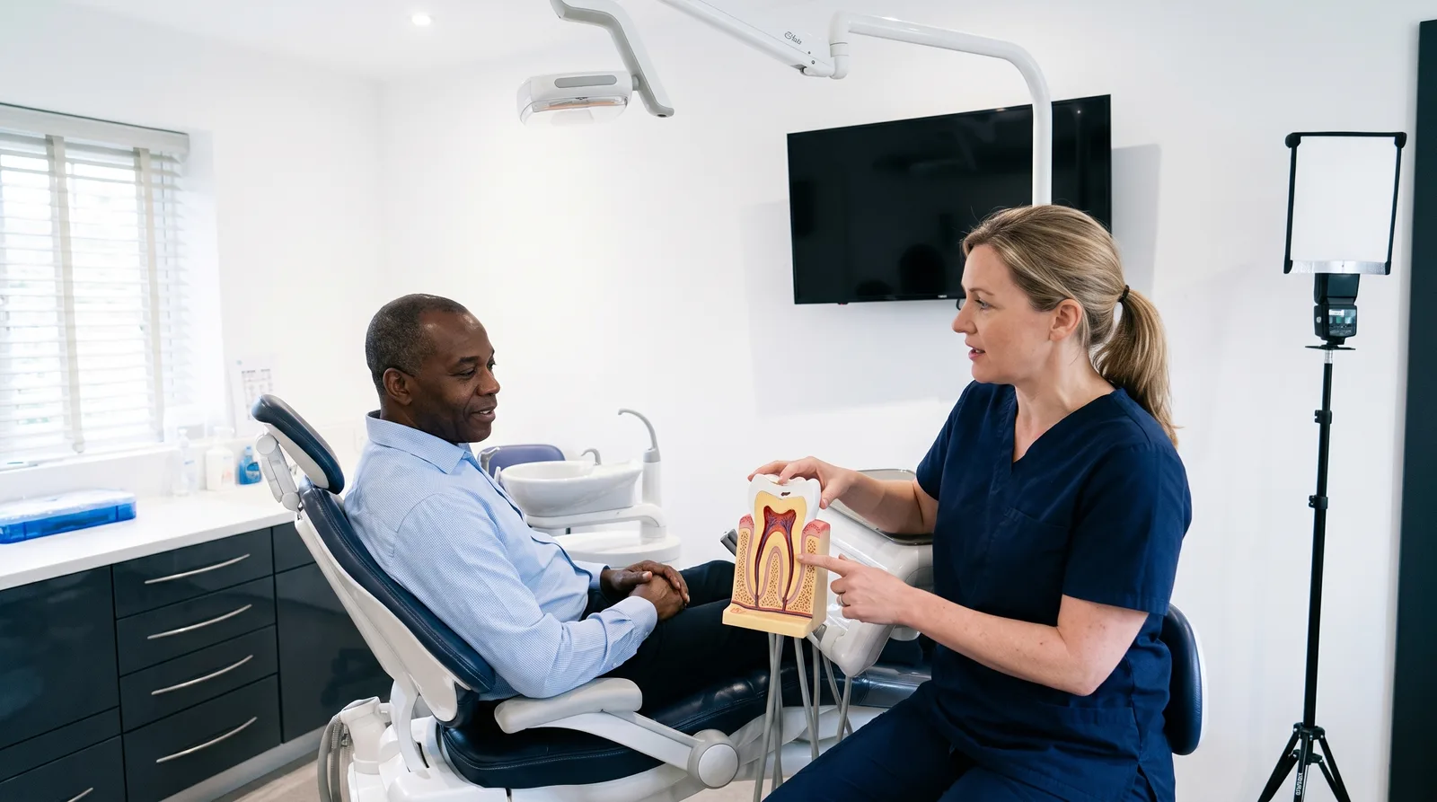A dentist in navy scrubs showing a seated patient a cross-section tooth model in a dental treatment room
