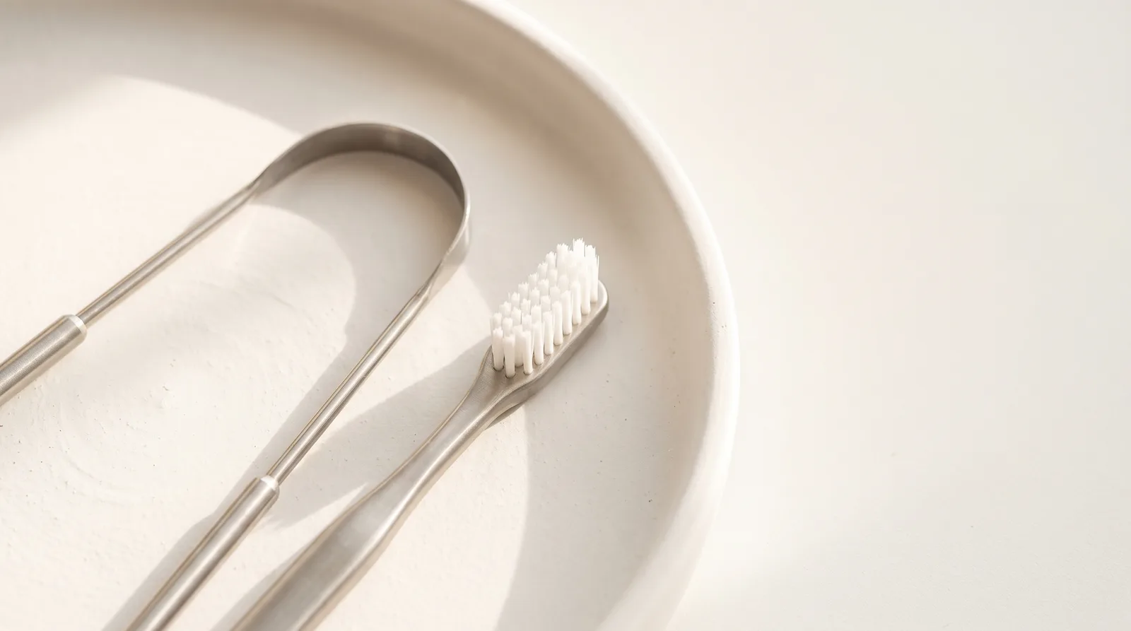 A stainless steel tongue scraper lying beside a toothbrush on a white surface