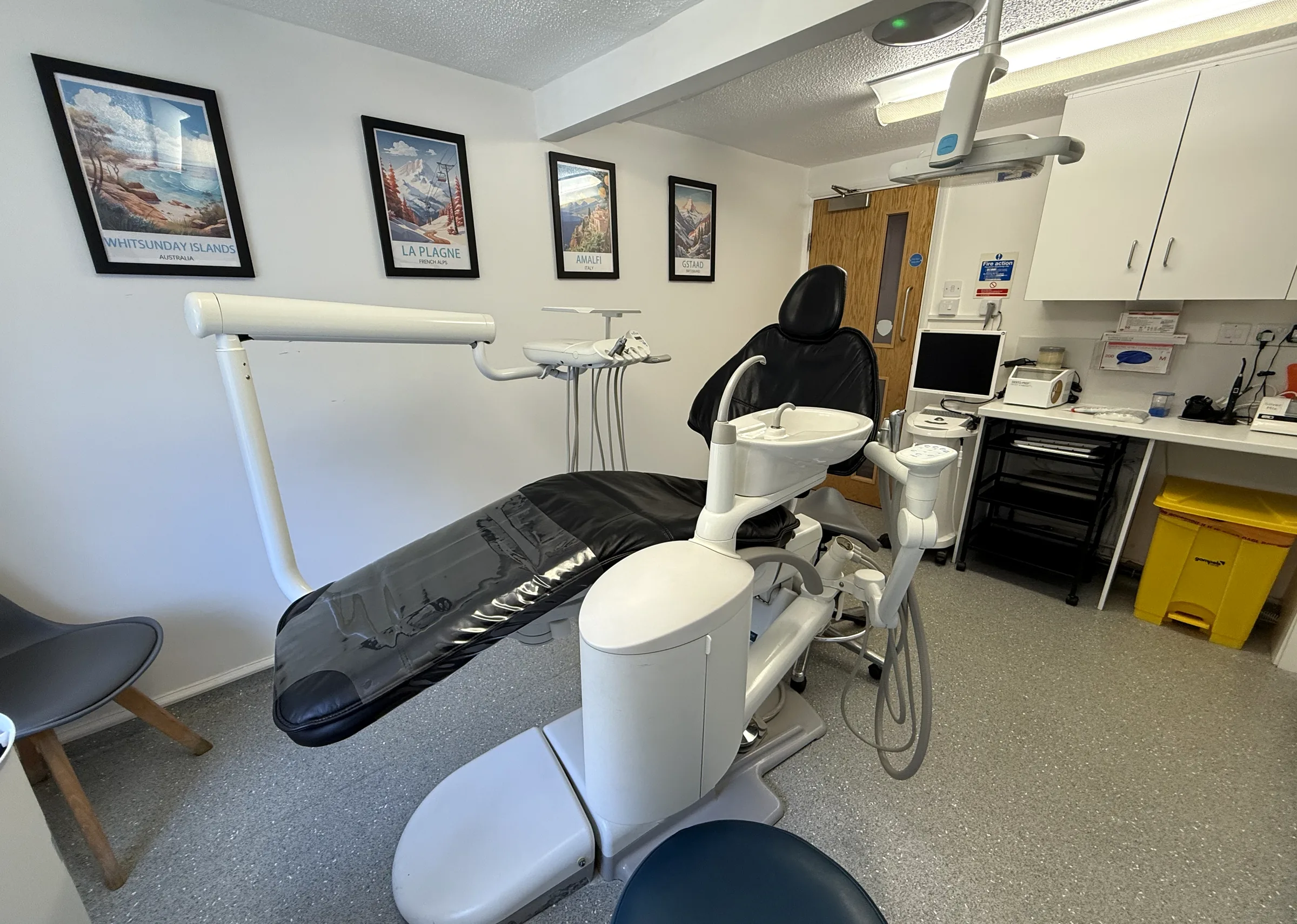 A treatment room at Earls Barton, with the chair, overhead light, worktop and framed travel prints